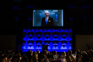 Governor Phil Murphy on screen speaking to gala guests in ballroom