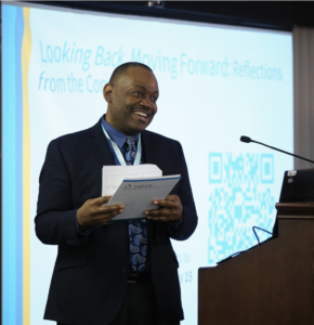 Speaker smiling in front of podium with presentation screen in background